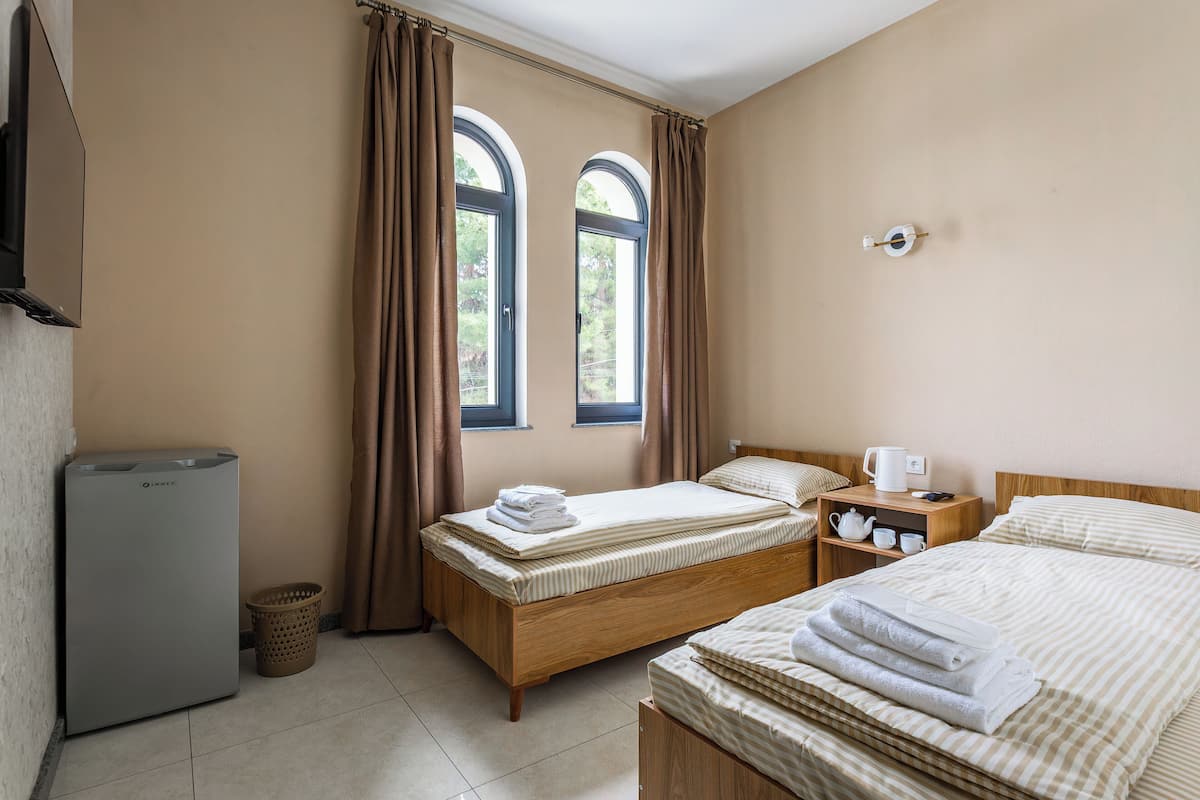 A hotel room with two beds, a small table with a kettle, a trash bin, and a window with curtains.