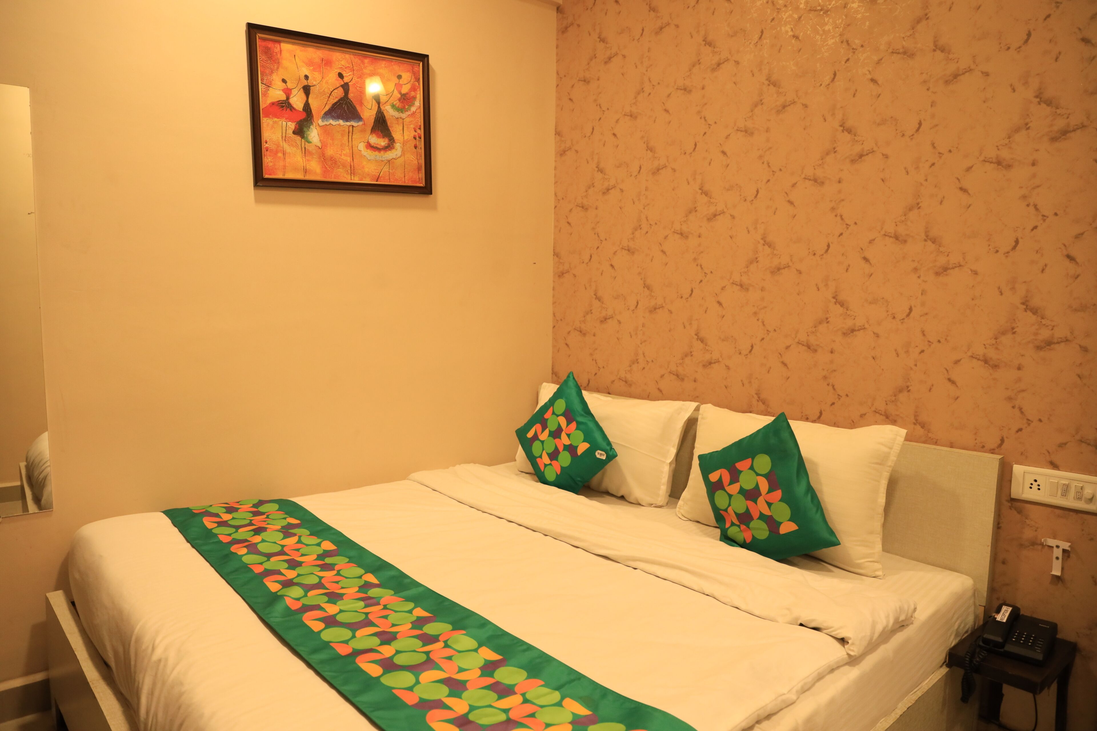 A hotel room with a bed, pillows, a blanket, a painting, a nightstand, and a phone.