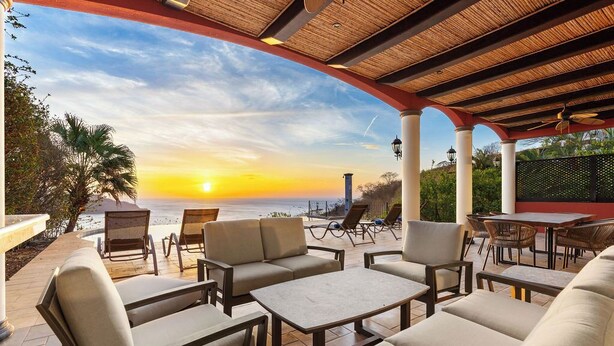 An outdoor seating area with a view of the sunset over the ocean, covered patio, and lounge chairs.