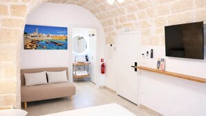 Ein Zimmer mit einem Steingewölbe, einer Couch, einem Fernseher und einem Badezimmer mit Spiegel, sowie ein Gemälde mit einer Küstenansicht.