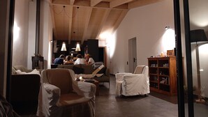 Un salon chaleureux avec un plafond haut, des poutres en bois et une cheminée. On y trouve plusieurs fauteuils et un canapé, ainsi qu’une bibliothèque et une lampe sur une table d’appoint.