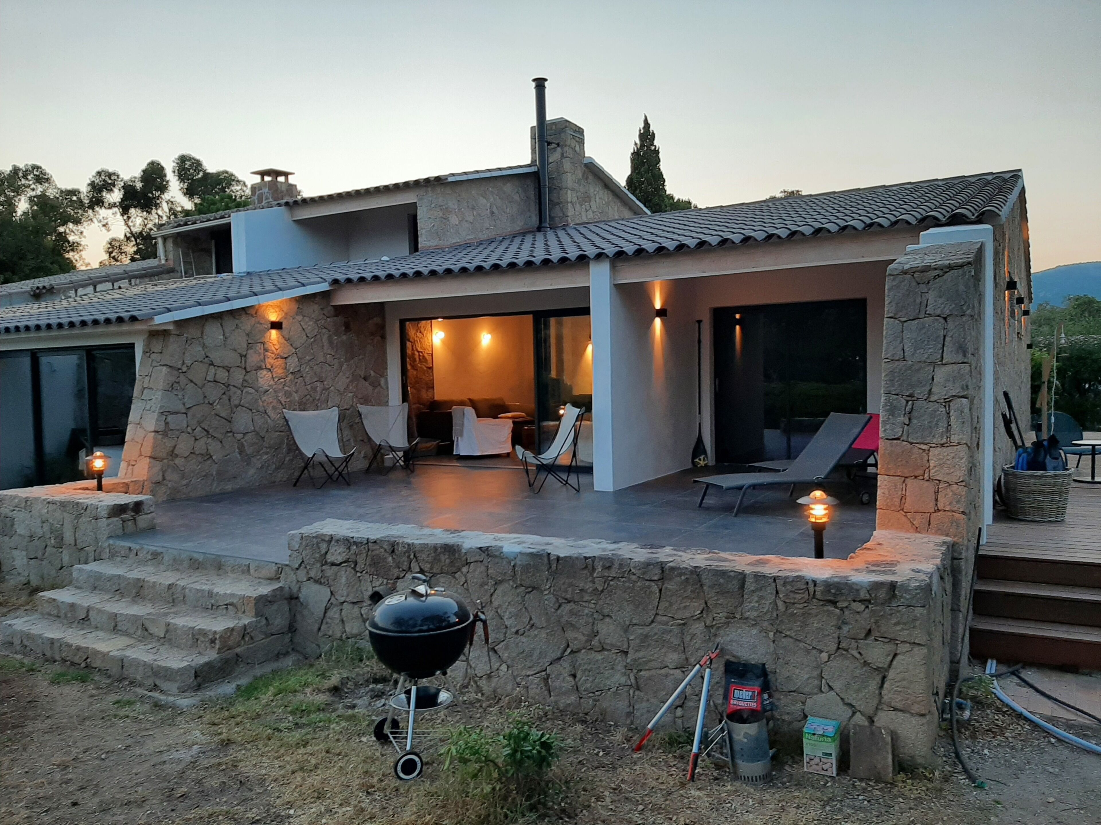 Ein modernes Haus mit Steinaußenwand, einer überdachten Terrasse mit Sitzgelegenheit und einem Grill.