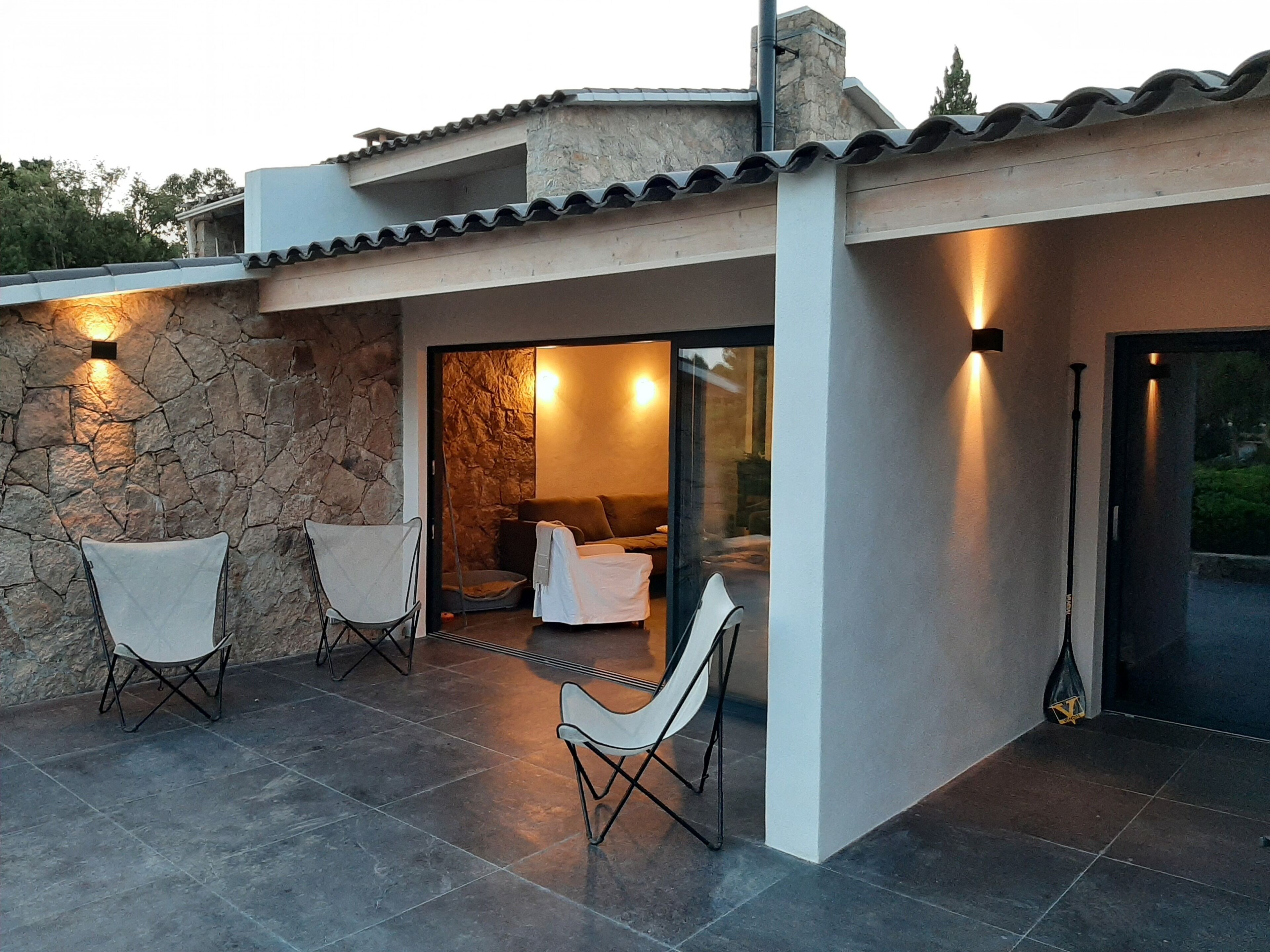 A modern house with a stone exterior, a patio with chairs, and a glass door leading to an interior space.