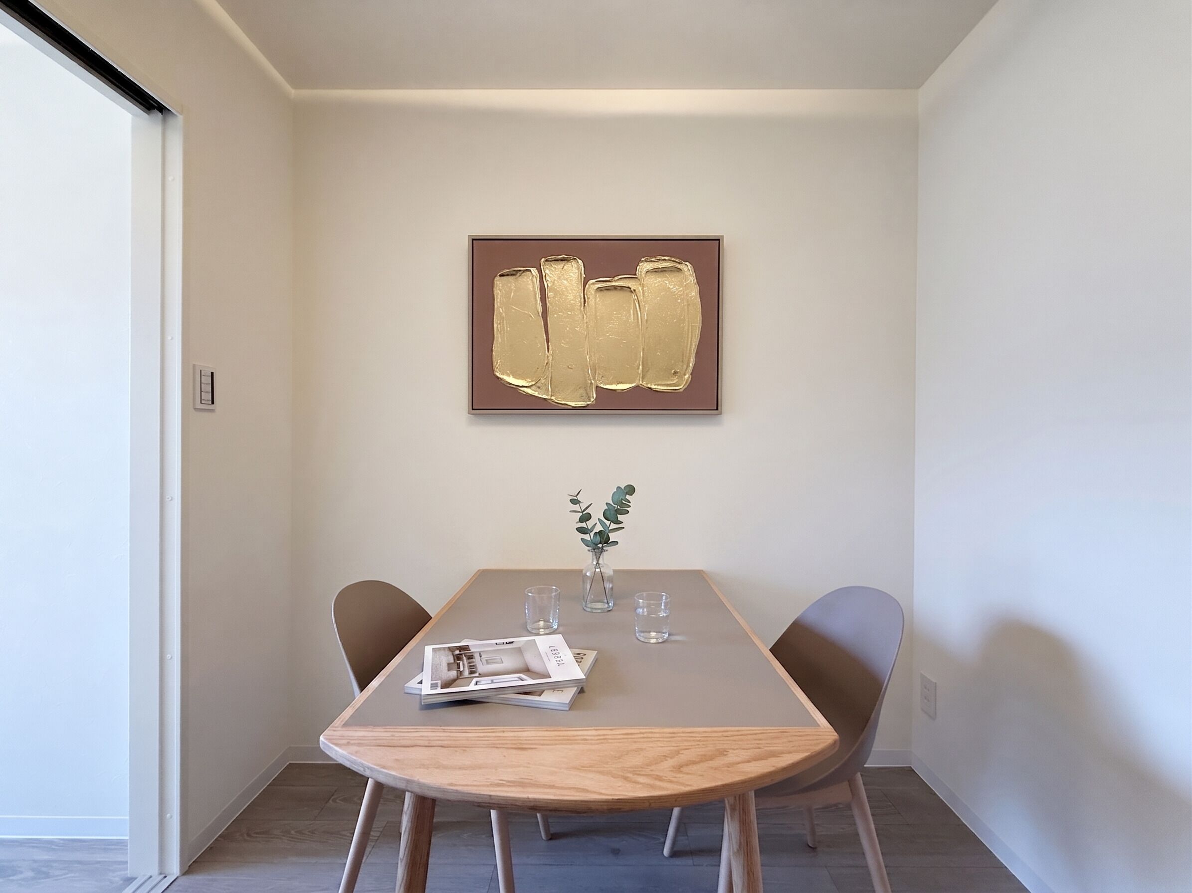 Een moderne eethoek met een houten tafel, twee stoelen, een vaas met plant en een lijst met een abstract schilderij aan de muur.