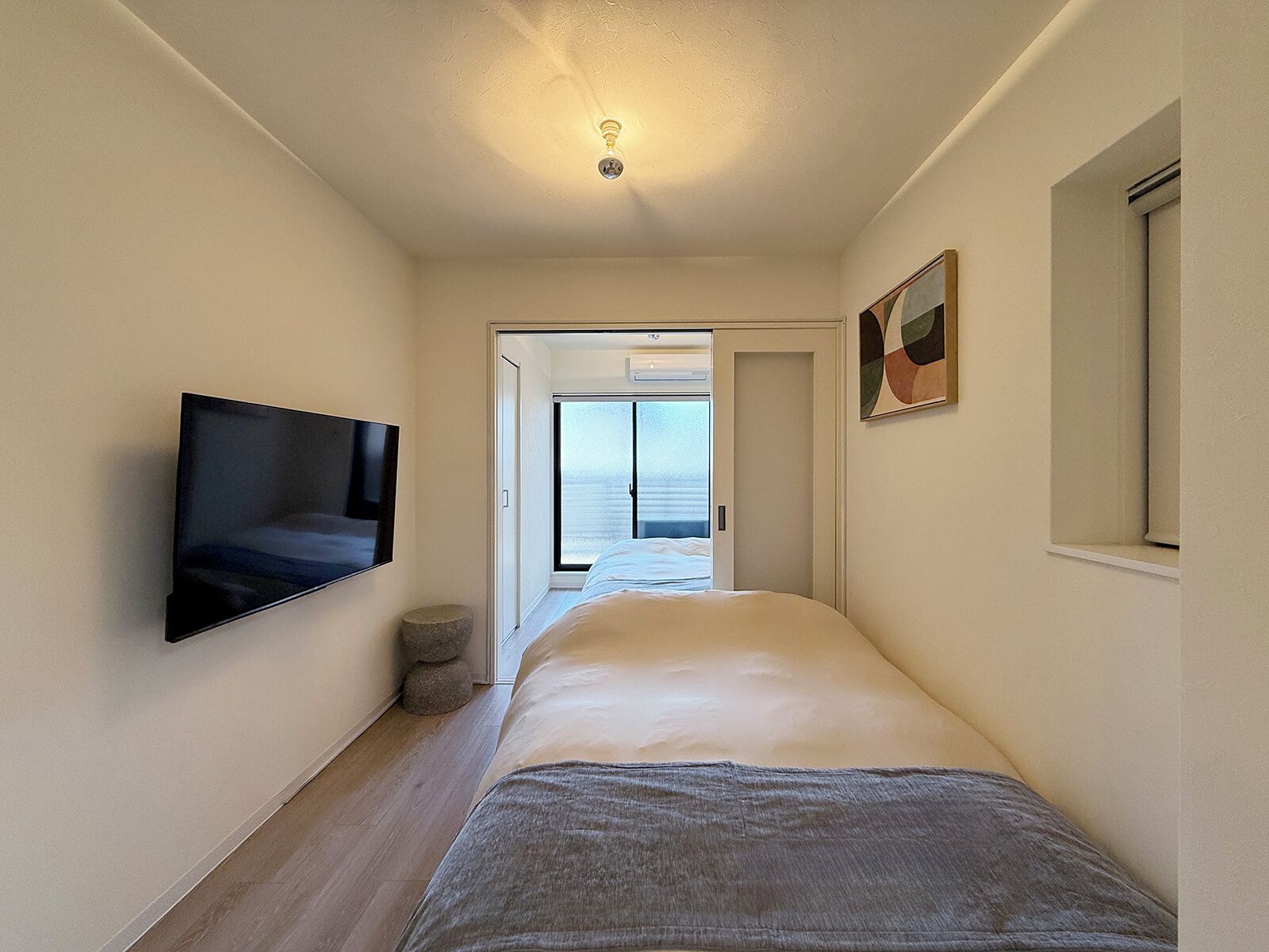 A narrow hotel corridor with a bed, a mounted TV, and a door leading to a balcony.