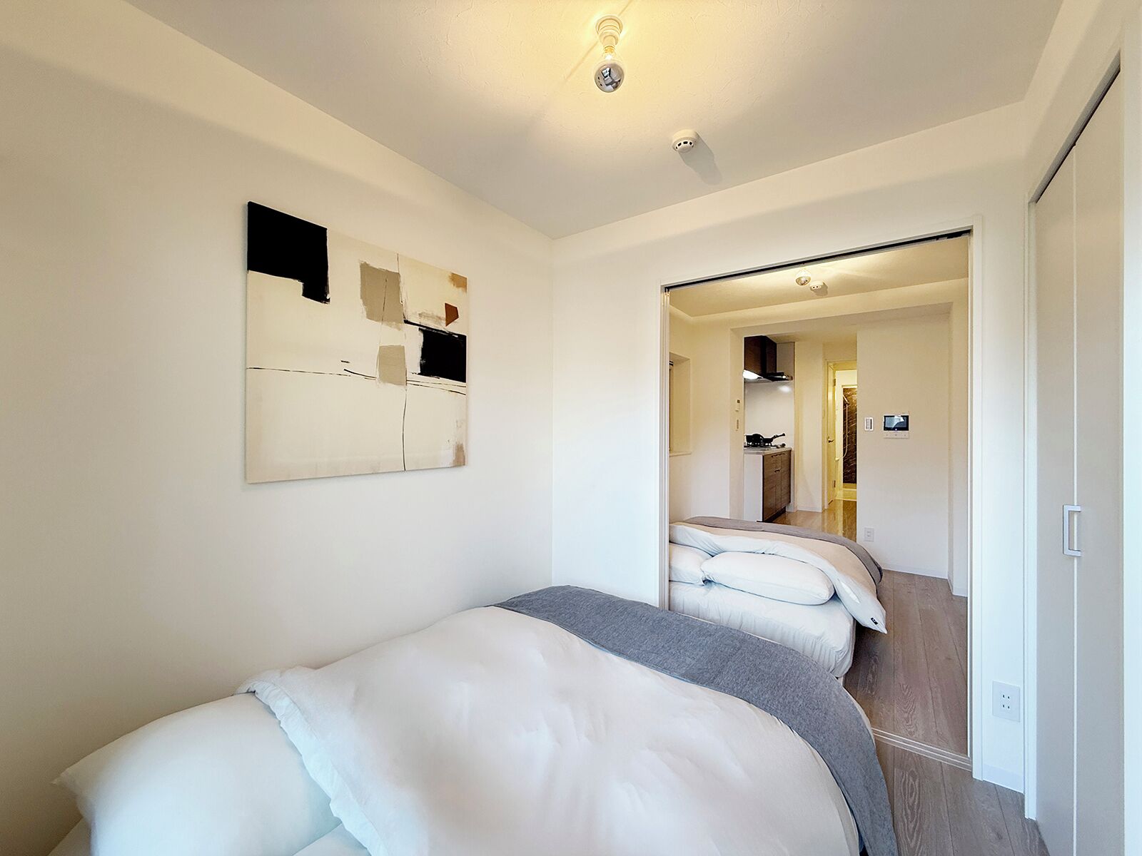 A bedroom with a bed, a painting, and a hallway leading to a kitchen.