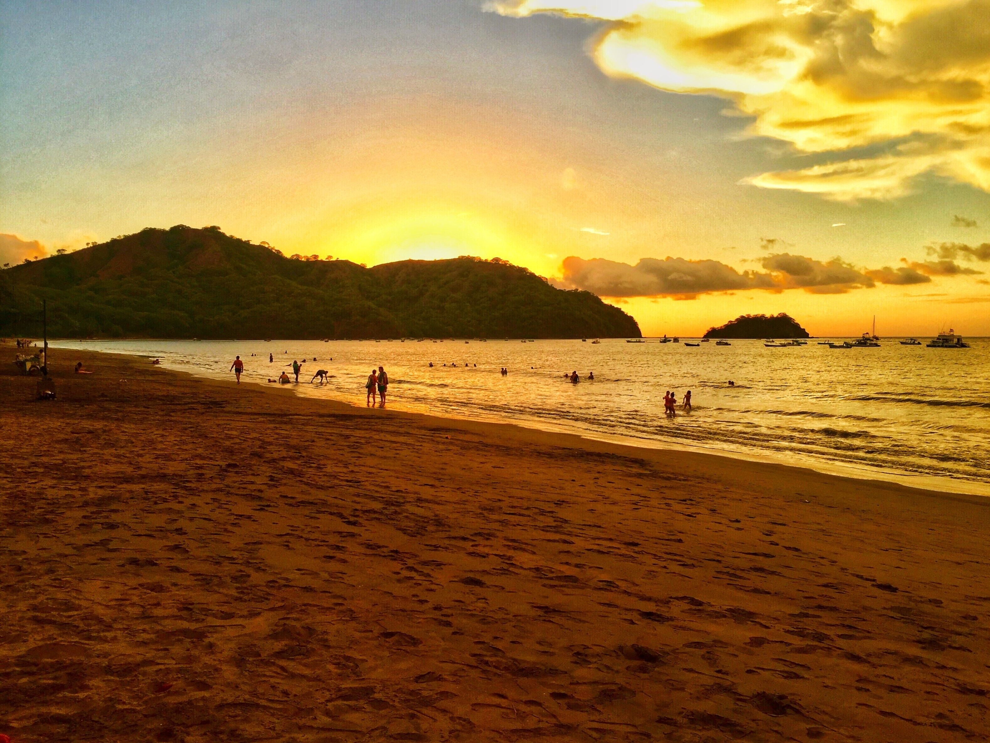 Una playa con gente caminando y nadando, barcos en el agua y un atardecer de fondo.