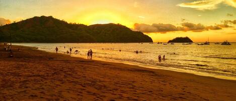 Una playa con gente caminando y nadando, barcos en el agua y un atardecer de fondo.