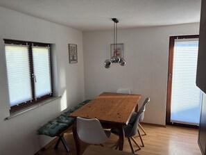A dining area with a wooden table, white chairs, a bench with a leaf pattern, and a ceiling light fixture.