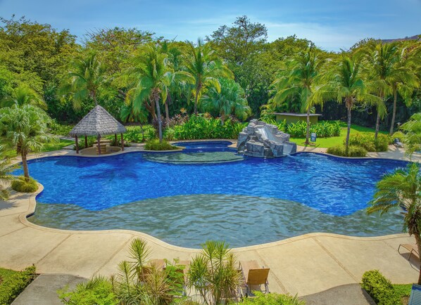 A large outdoor swimming pool with a rock feature, surrounded by lush greenery and palm trees.