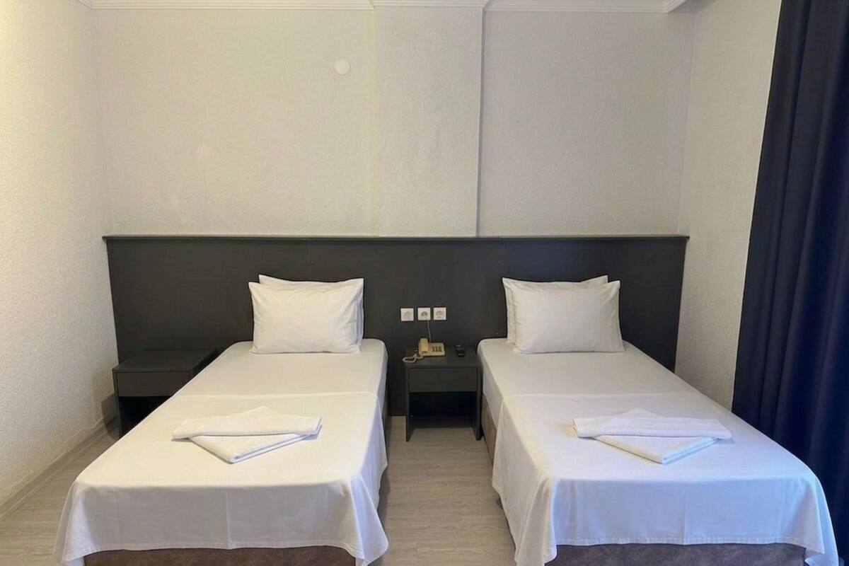 A hotel room with two single beds, a dark headboard, a small bedside table with a phone, and a dark curtain on the right.