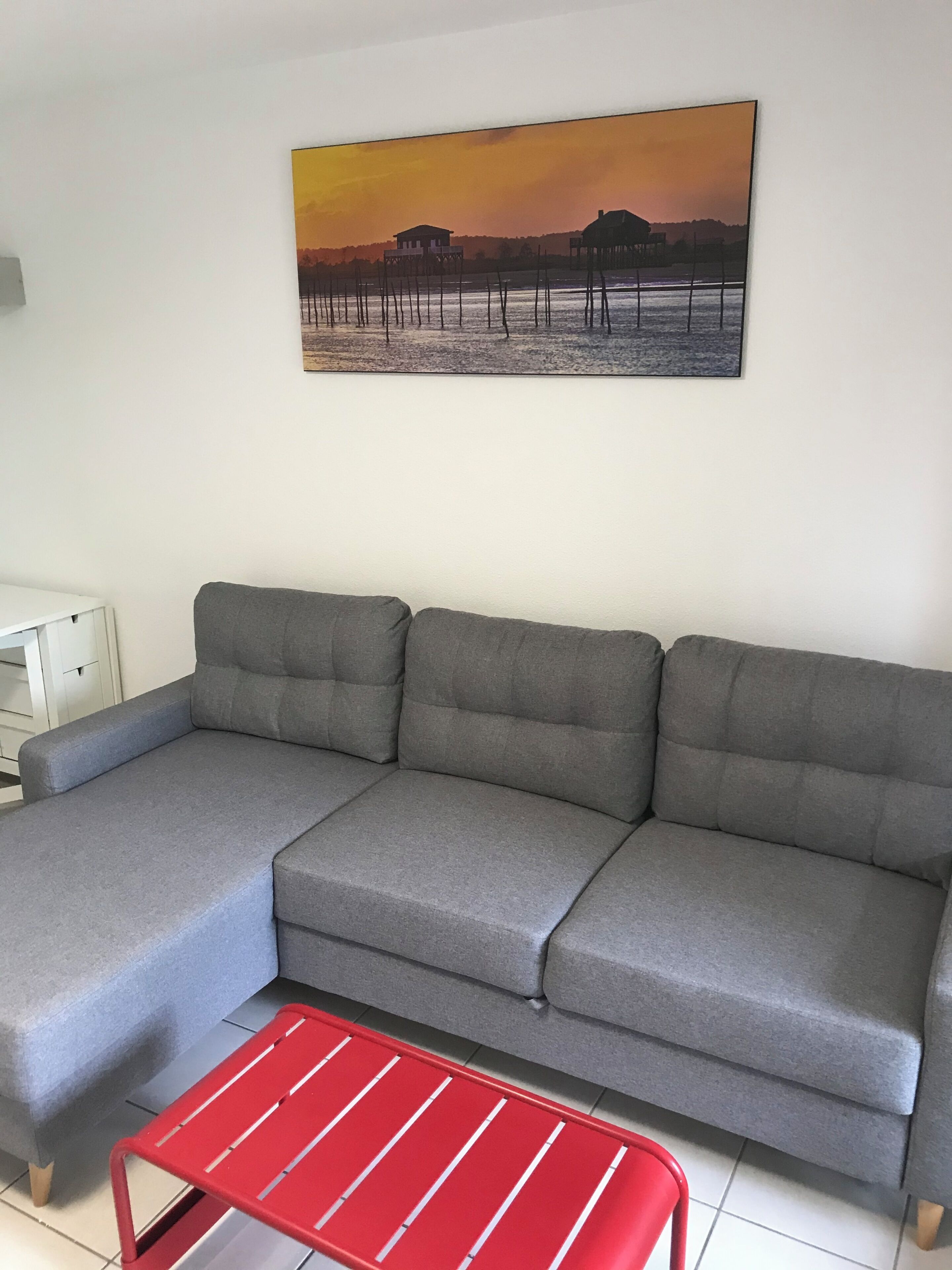 A grey sectional sofa, a red metal table, and a sunset landscape painting on the wall.