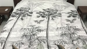 Una cama con cabecera de madera oscura, una colcha blanca con estampado de hojas tropicales y pájaros, y dos mesitas de noche, con un control remoto en una de ellas.