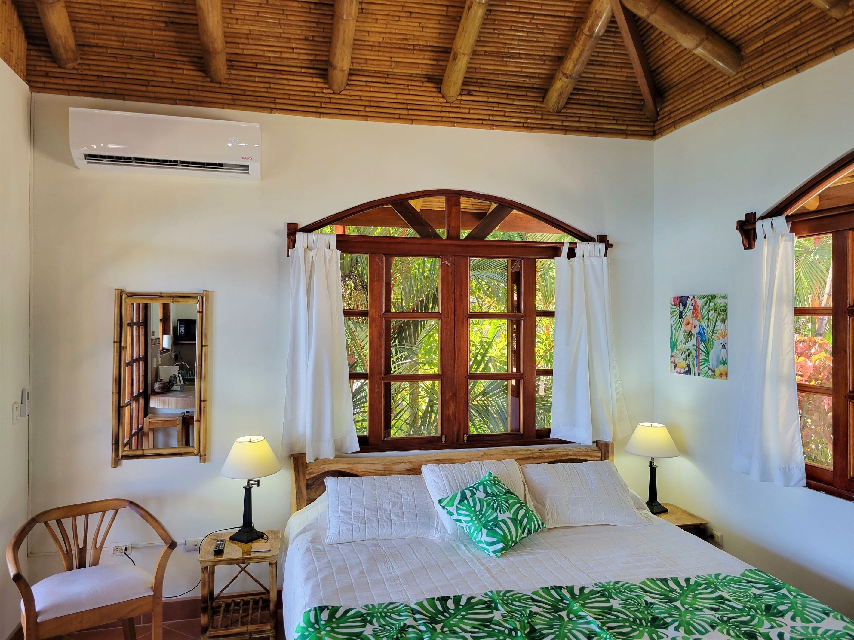 A bedroom with a wooden ceiling, a large window with curtains, a bed with white bedding, a nightstand with a lamp, and a chair.