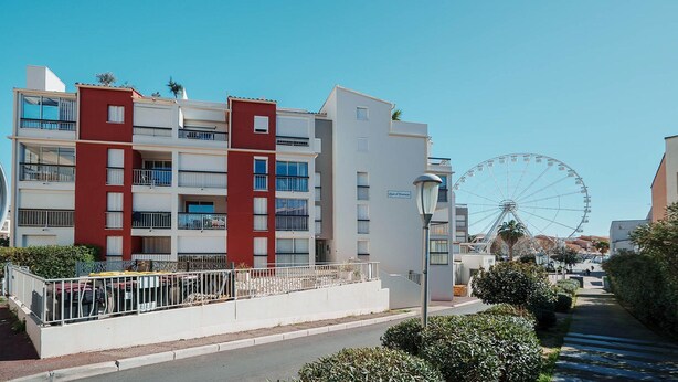 A multi-story building with balconies, a Ferris wheel in the background, and a street with a sidewalk and greenery. - Sophisticated Retreat Near the Sea, Agde (Agde)