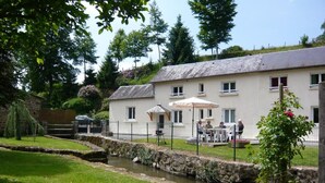 A white building with a stone wall, a small stream, and outdoor seating with umbrellas. - The Monthorin Valley (La Bazouge-du-Désert)