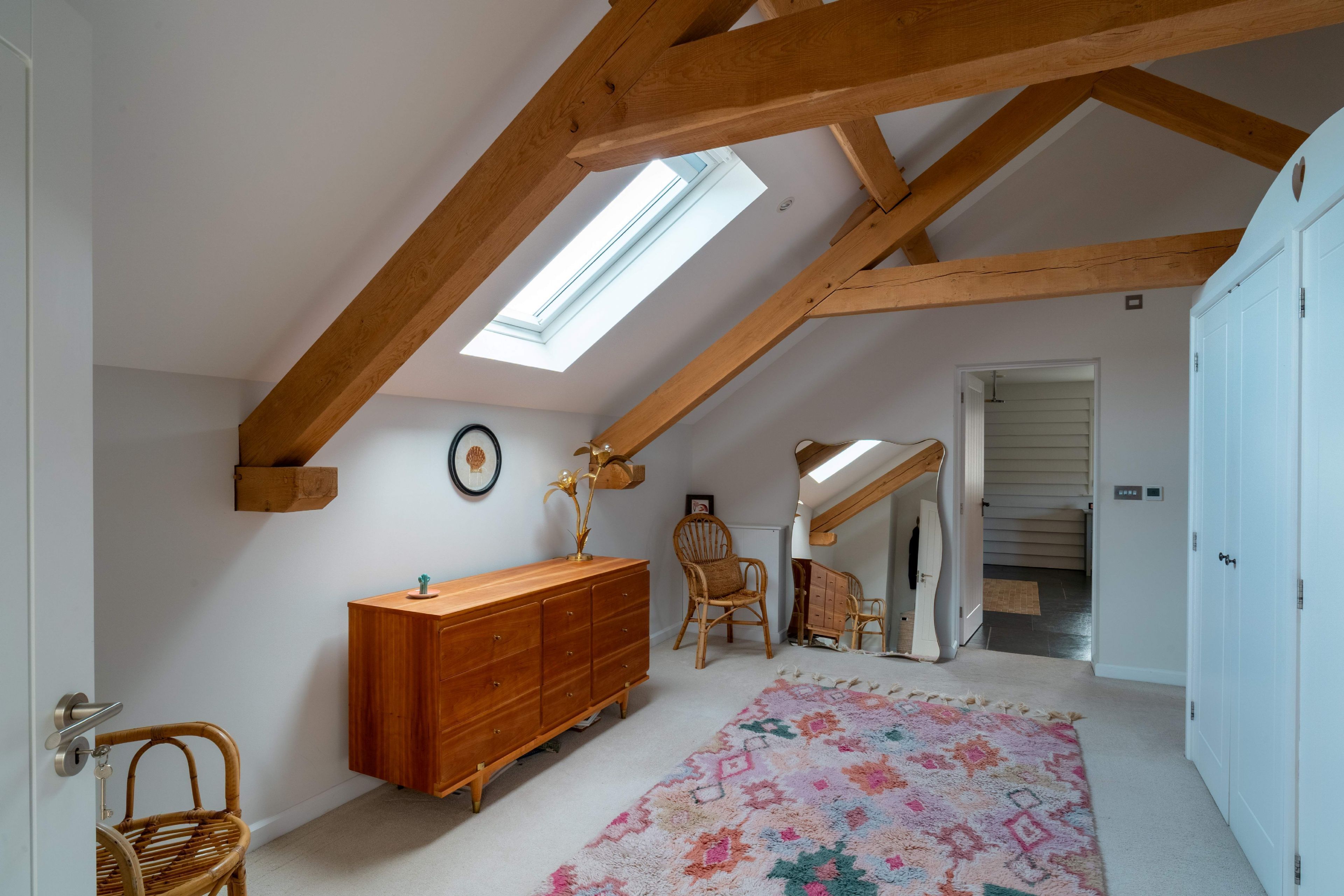 A room with a sloped ceiling, wooden beams, a large window, a wooden dresser, a wicker chair, a patterned rug, and a door leading to another room.