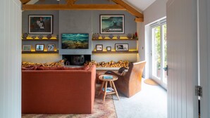 A living room with a fireplace, sofas, a coffee table, and a bookshelf with framed pictures and decorative items. - Some homes are made for holidays,  Surfers Roost is made for time well spent together. (Croyde)