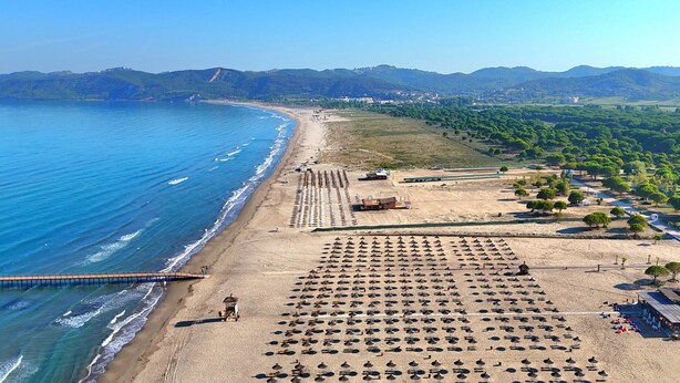 Une vue aérienne d’une plage bordée de rangées de parasols et de chaises longues, avec une jetée qui s’avance dans la mer, et un paysage en arrière-plan composé de collines et de bâtiments.