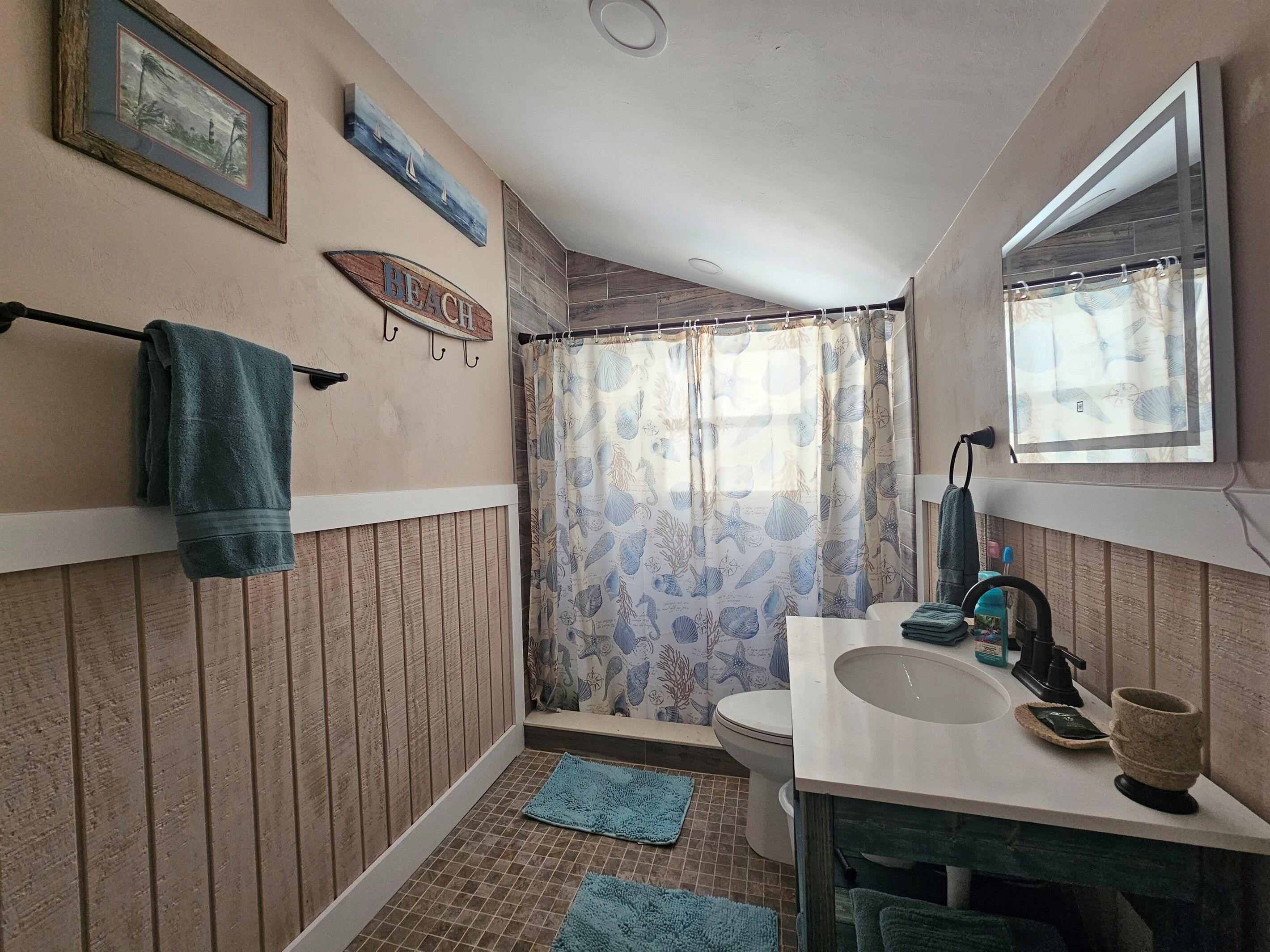 Ein Badezimmer mit Holzwandverkleidung, Toilette, Waschbecken mit Spiegel und einer Duschvorhang mit Meeresmotiven.