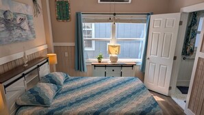 A bedroom with a bed, bedside tables, a window, a door, and a wall decoration. - Seafoam Cottage - Beauty in the Heart of Cedar Key (Cedar Key)
