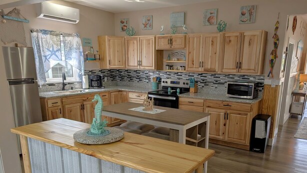 A modern kitchen with wooden cabinets, a stainless steel refrigerator, a microwave, and a dining area with a table and chairs. - Seafoam Cottage - Beauty in the Heart of Cedar Key (Cedar Key)