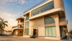 A modern building with a sign reading 'The Royal Palace.' It features a curved facade, large windows, and a prominent entrance with steps. - Travinities Hotel - The Royal Palace (Indore)