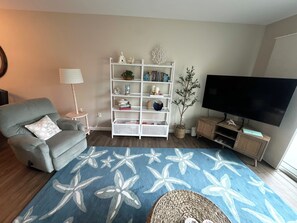 Ein Wohnzimmer mit grauem Sessel, einem weißen Bücherregal, einem blauen Teppich, einem Fernseher und einem kleinen Tisch.