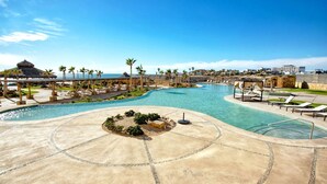 A resort pool area with a curved pool, lounge chairs, palm trees, and a thatched-roof structure. - Beachfront 1BR High-floor Panoramic Views (Puerto Peñasco)