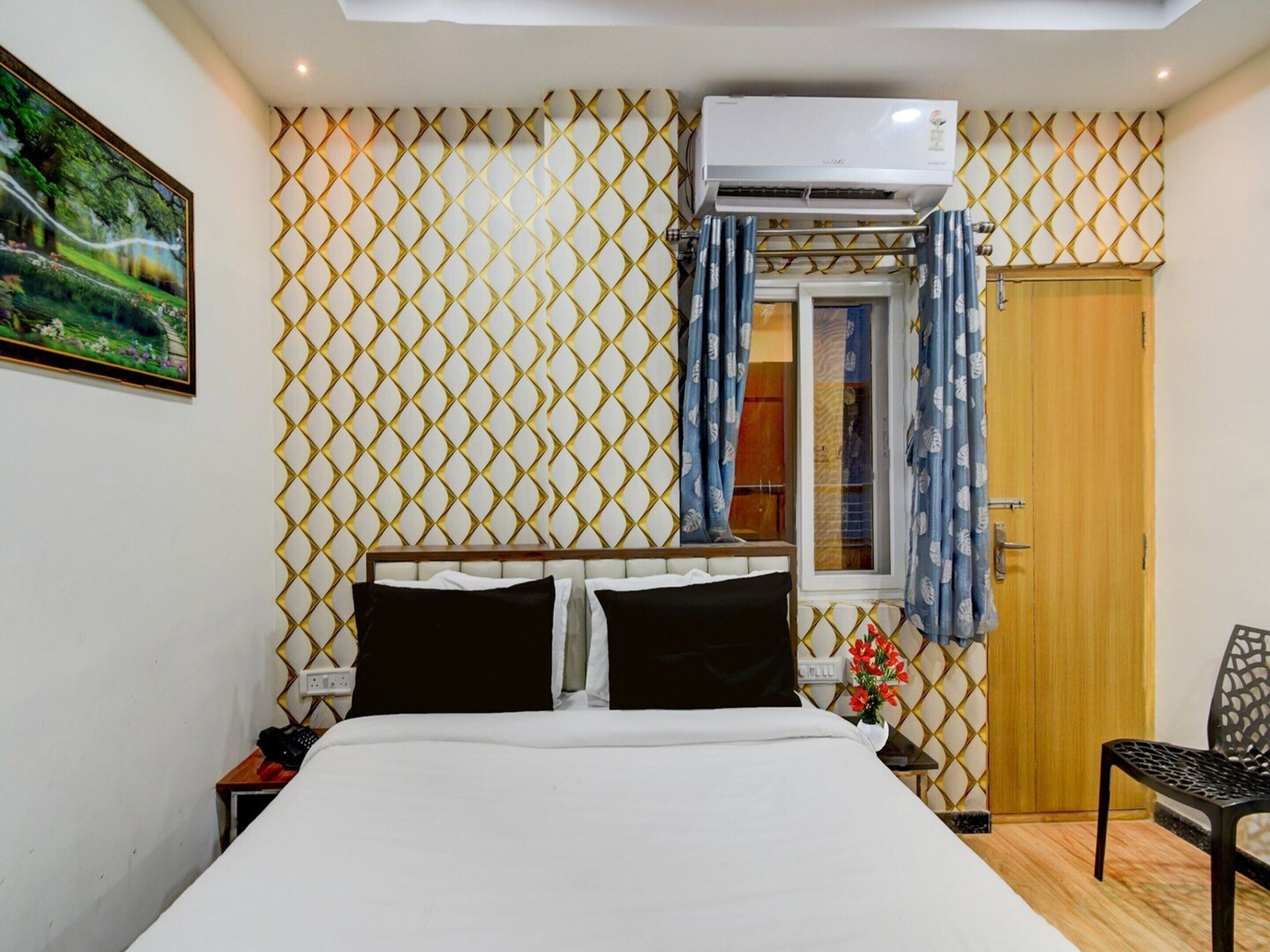 Una habitación de hotel con cama, mesita de noche, silla, aire acondicionado, ventana con cortinas y puerta.