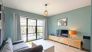 A modern living room with a sofa, a TV on a wooden console, a large window with curtains, and a hanging light fixture. - Eldon Terrace - Luxury 1 Bedroom Property (York)