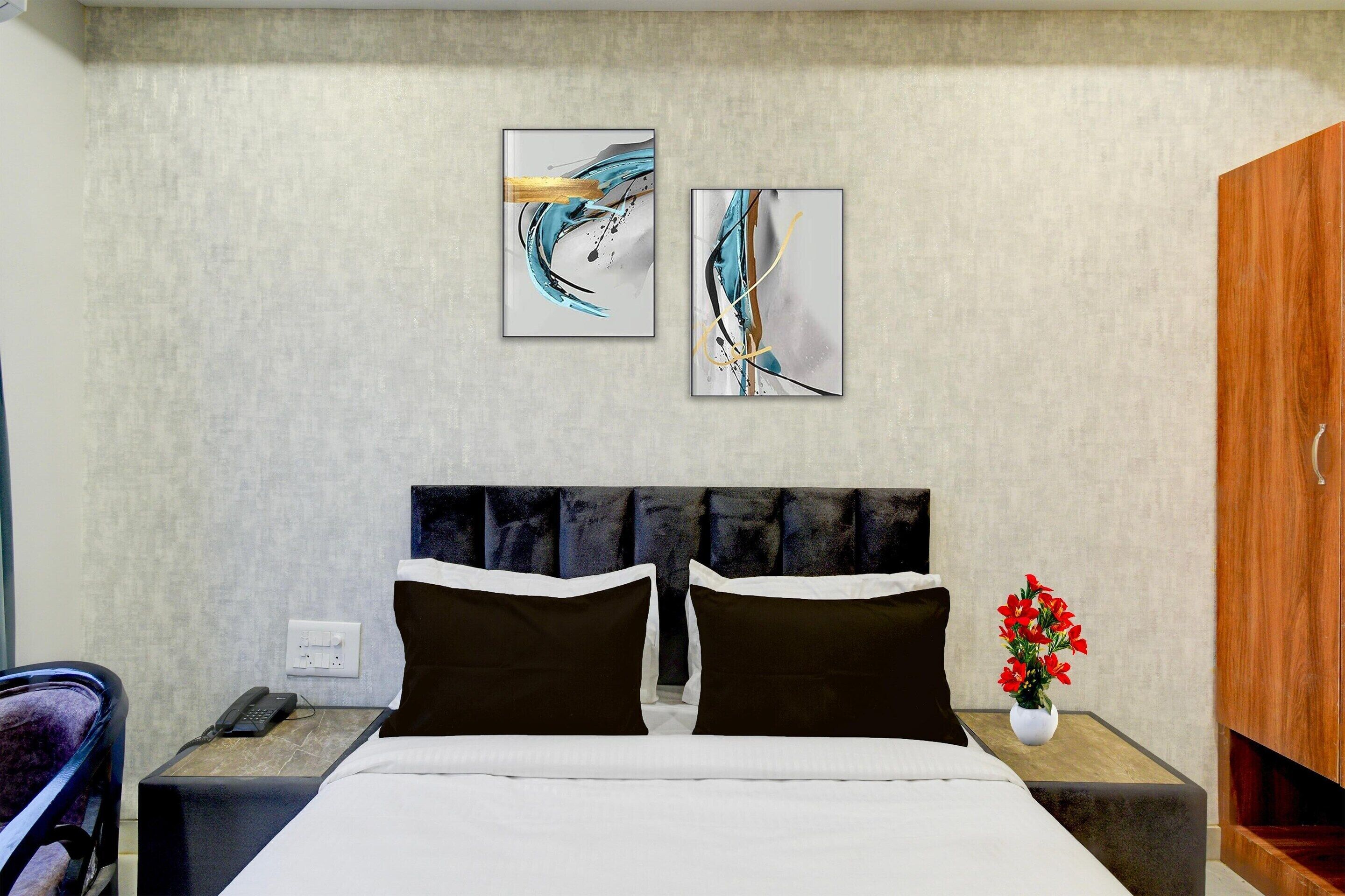 Una habitación de hotel con cama, mesita de noche, teléfono, armario y dos cuadros abstractos en la pared.