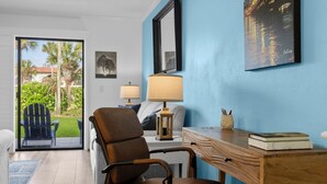 Une pièce donnant sur un jardin, avec un bureau en bois, une chaise, une lampe et un tableau encadré au mur.