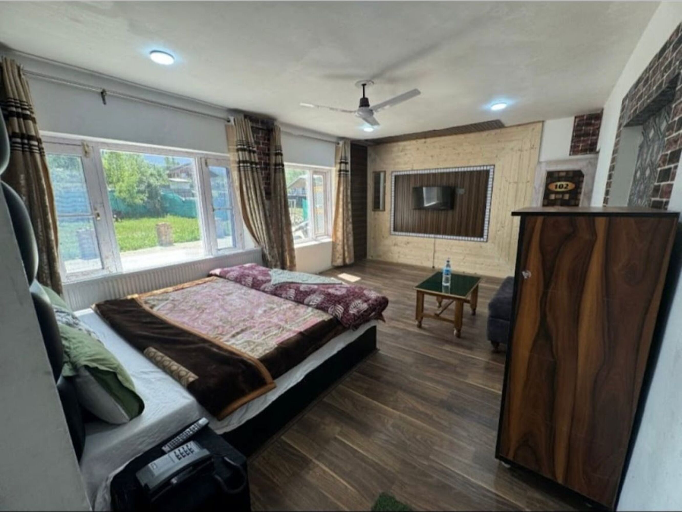 A bedroom with a bed, a TV mounted on the wall, a wooden wardrobe, and a small table.