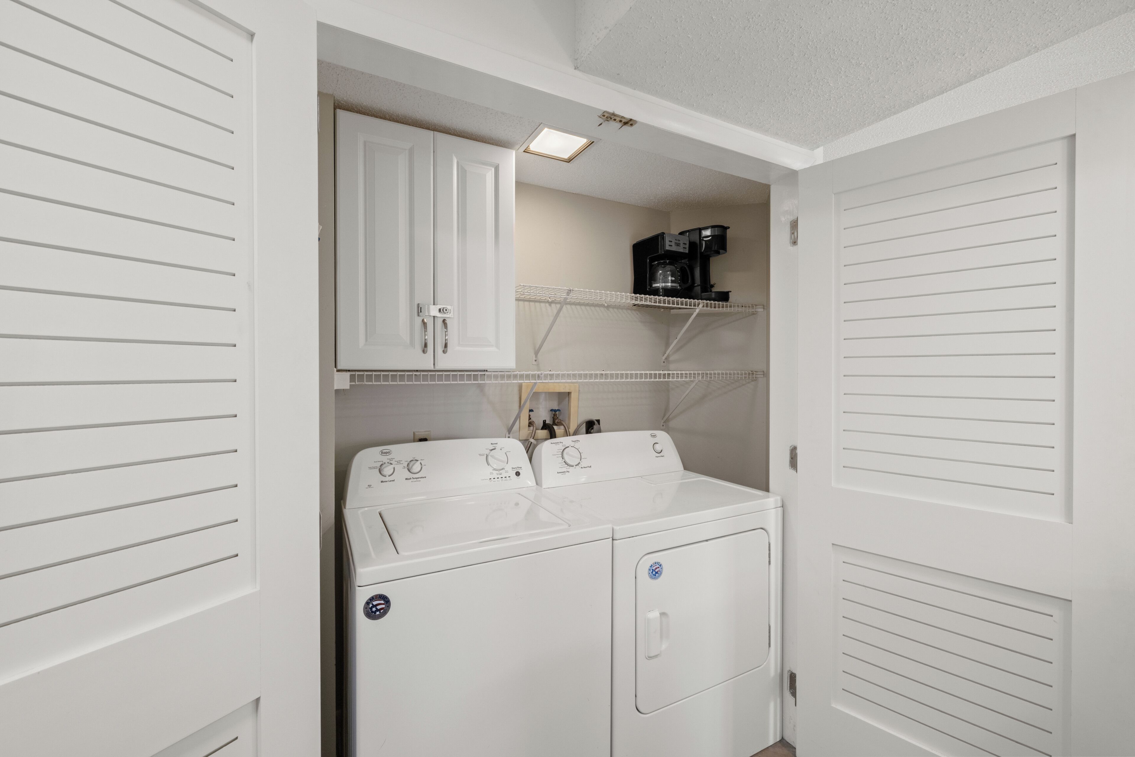 Une buanderie équipée d’un lave-linge et d’un sèche-linge, avec une machine à café posée sur une étagère et un meuble de rangement au-dessus du sèche-linge.