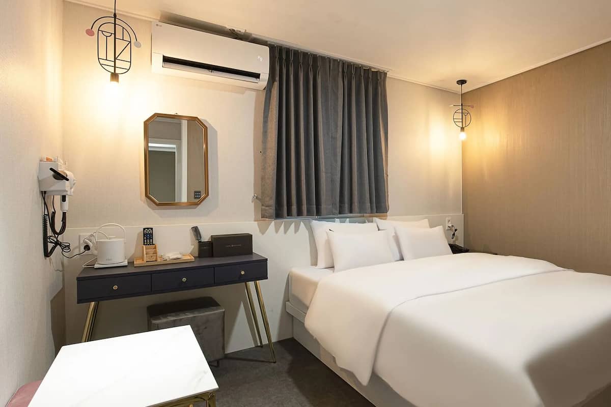 A hotel room with a bed, bedside table, mirror, air conditioning unit, and a window with curtains.