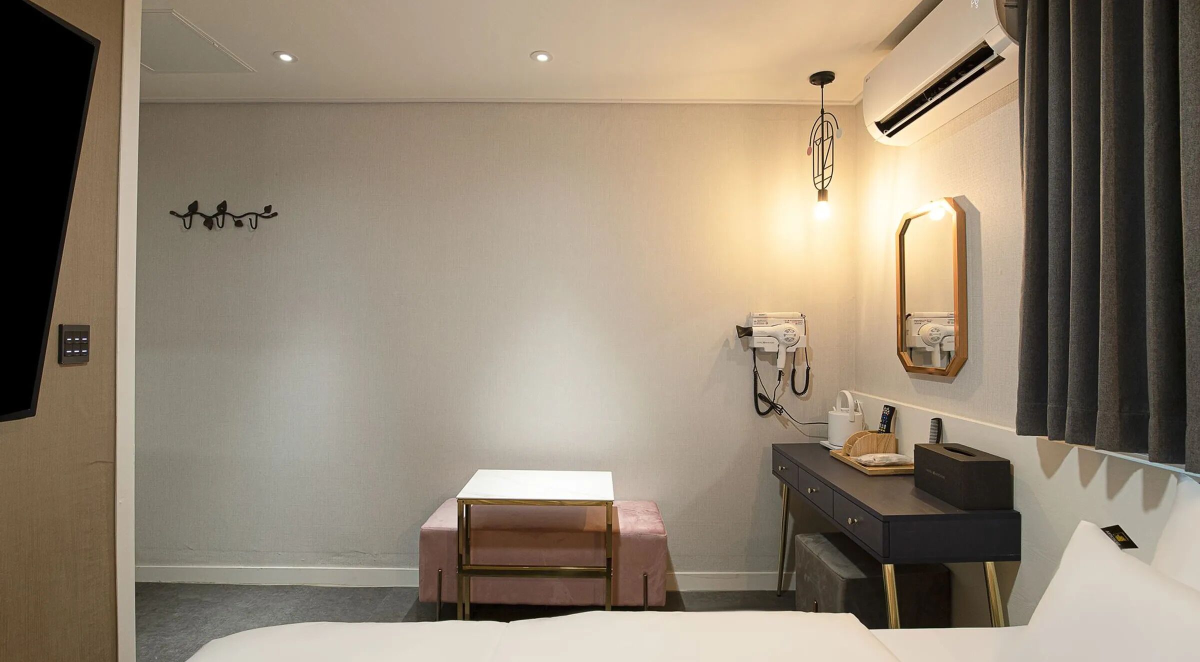A hotel room with a bed, a small table, a vanity with a mirror, a hairdryer, and an air conditioning unit.
