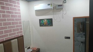 A bedroom with a bed, a bedside table, a wall-mounted air conditioner, a small painting, and a bathroom visible through an open door. - ; THE PERFECT HAVEN FOR YOUR COMFORT'' A PEASEFUL RETREAT , (Vrindavan)