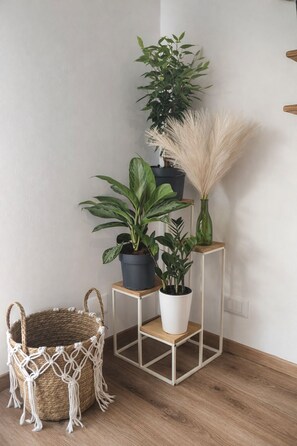 Eine Ecke mit einem gewebten Korb, einem Wandbehang aus Makramee und einem weißen Metallgestell mit drei Blumentöpfen.