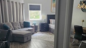 A modern living room with a grey sofa, a small side table, a lamp, a bed with a dark headboard, and a window with blinds. - Luxe waterfront cottage with a private dome (Whitchurch-Stouffville)