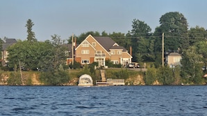 A large house with a prominent front porch and a small bridge leading to the water. - Luxe waterfront cottage with a private dome (Whitchurch-Stouffville)
