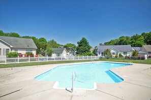 Piscina retangular com deck branco, cercada por uma grade branca, e casas ao fundo.