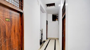 Um corredor estreito com uma porta de madeira identificada com o número '001' à esquerda, um chão de piso preto e branco e uma parede branca.