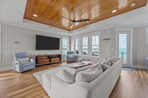 A spacious living room with a large gray sectional sofa, a wooden ceiling, a flat-screen TV, and a view of the ocean through large windows. - Observation Cove #217 (Topsail Beach)