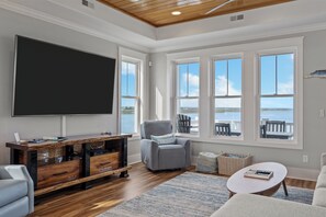 A spacious living room with a large flat-screen TV, wooden entertainment center, comfortable seating, large windows with a view of the water, and a ceiling with wooden beams. - Observation Cove #217 (Topsail Beach)