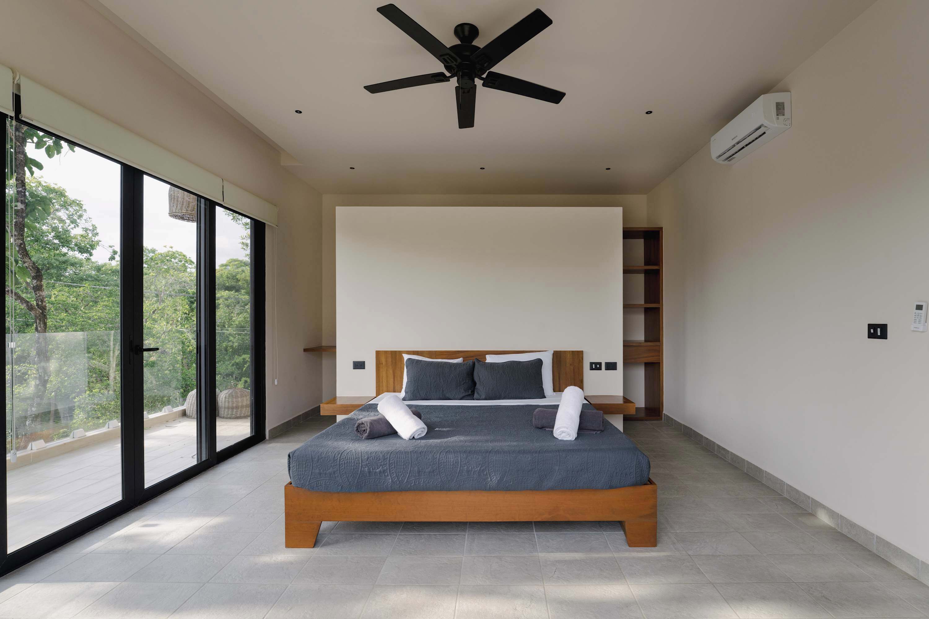 A modern bedroom with a large bed, a ceiling fan, and a view through large windows.