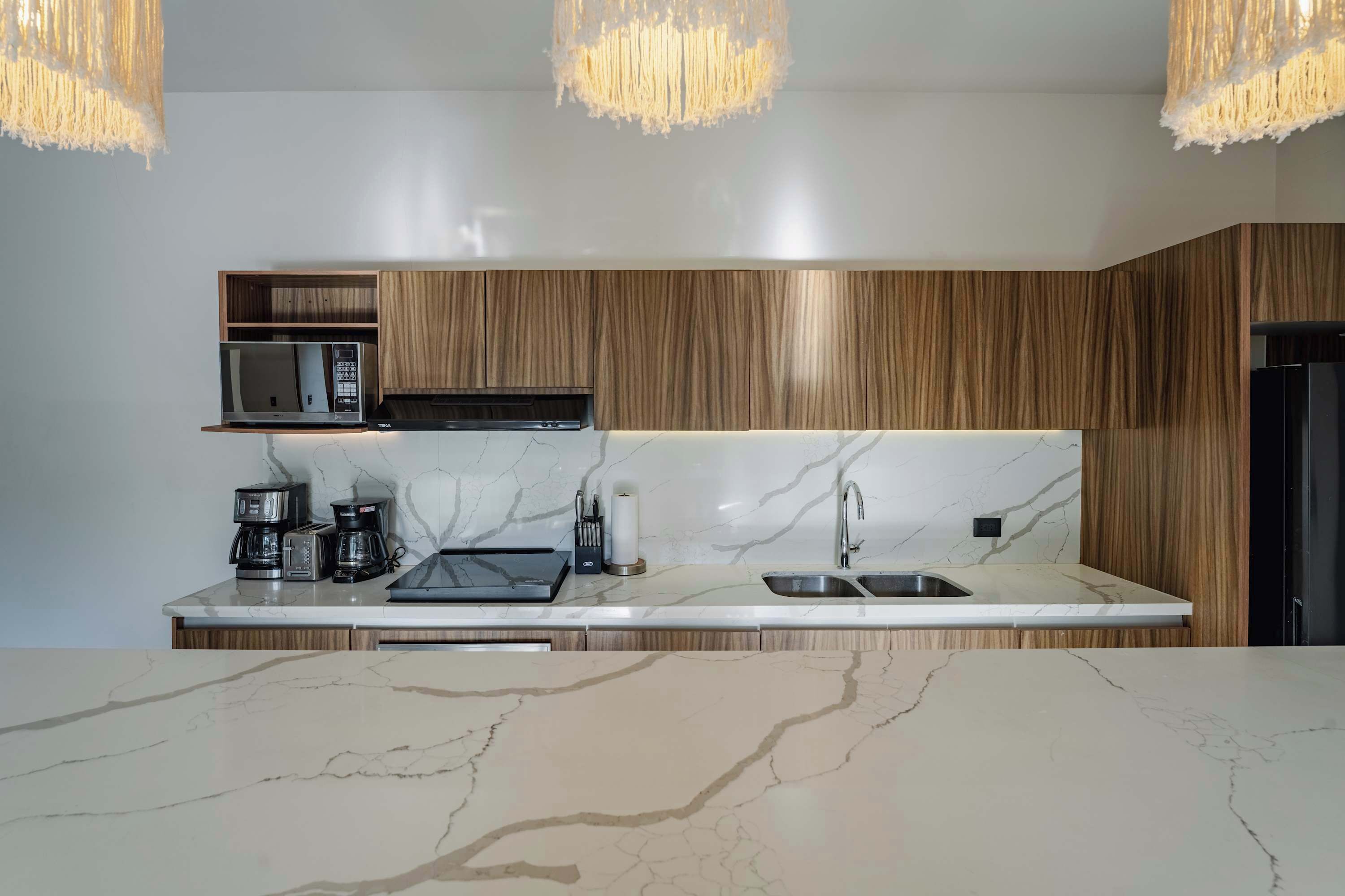 A modern kitchen with a marble countertop, wooden cabinets, a microwave, a coffee maker, and a sink.