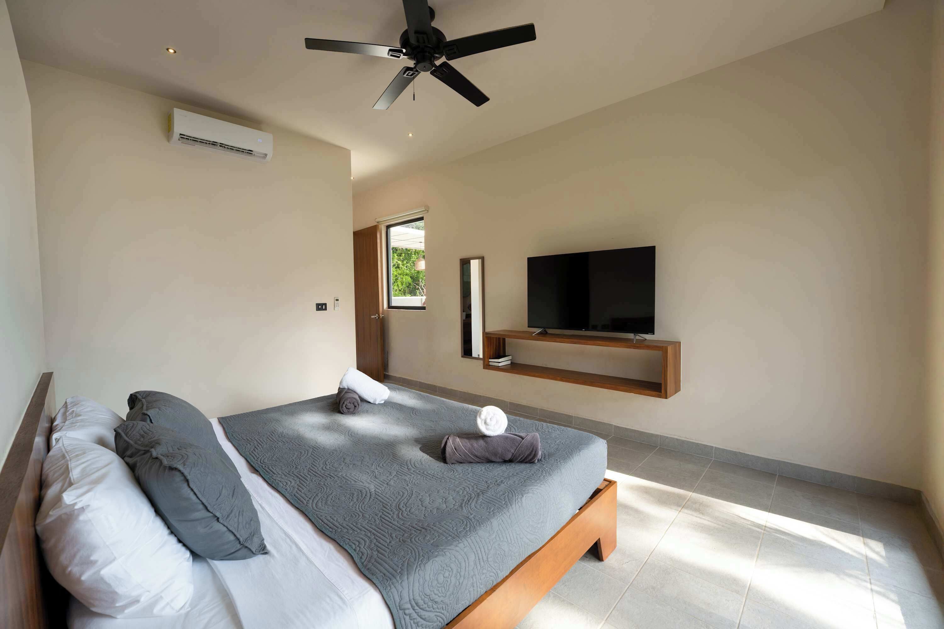 A bedroom with a bed, pillows, a ceiling fan, an air conditioner, a TV on a wooden shelf, and a window.