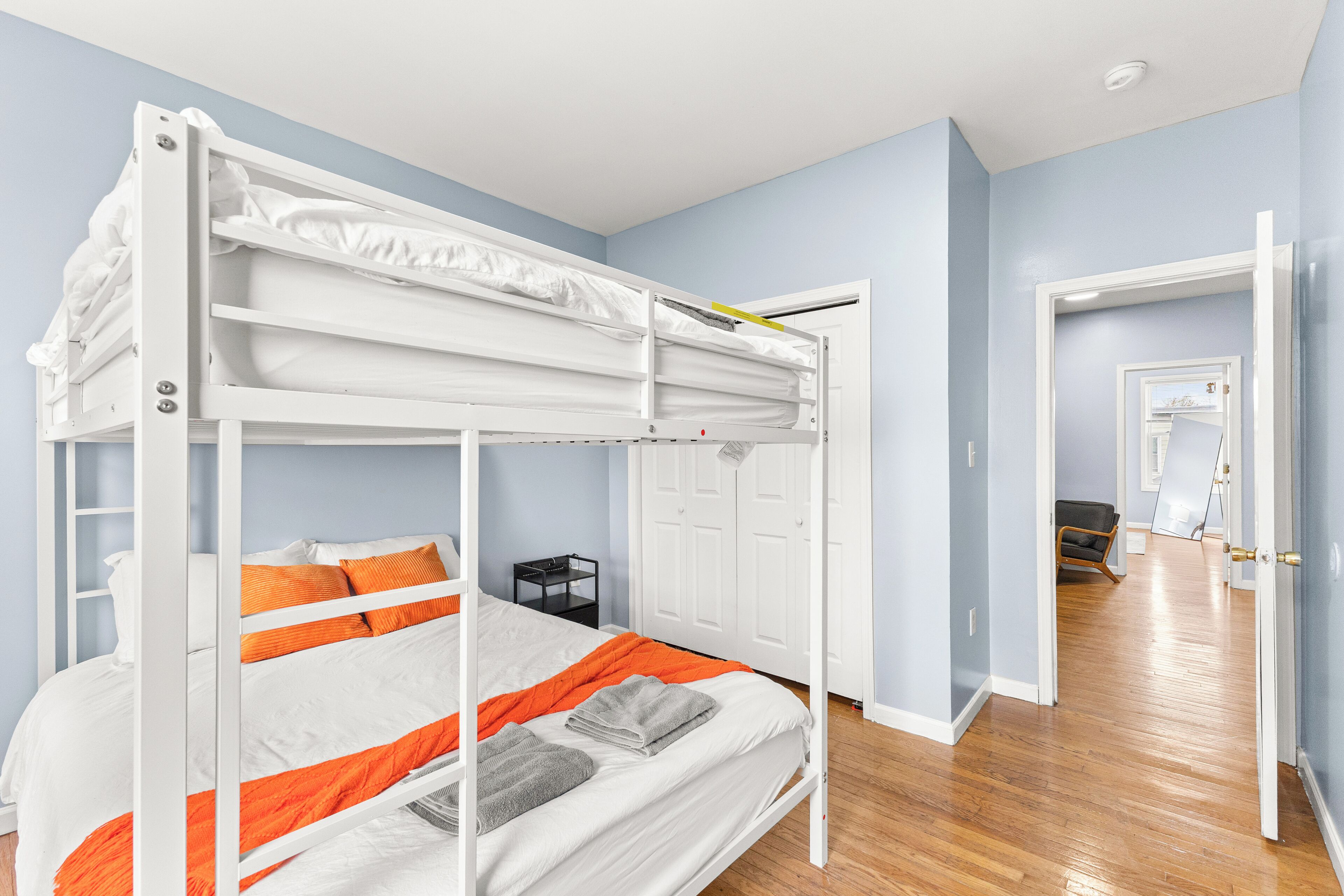 Ein Stockbett mit weißer und orangefarbener Bettwäsche, ein schwarzer Nachttisch und eine weiße Tür in einem Zimmer mit hellblauen Wänden und Holzboden.