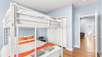 Ein Stockbett mit weißer und orangefarbener Bettwäsche, ein schwarzer Nachttisch und eine weiße Tür in einem Zimmer mit hellblauen Wänden und Holzboden.